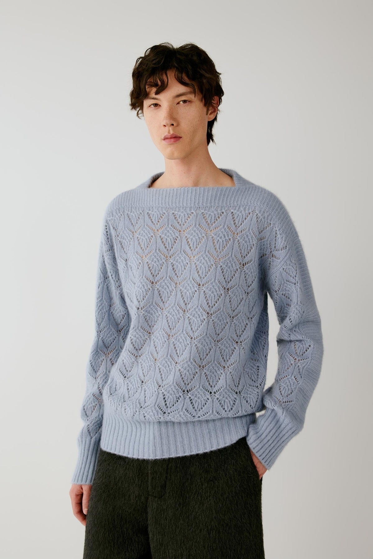 A man wearing a light blue knit sweater with a textured geometric pattern and dark charcoal pants, standing against a plain light gray background.