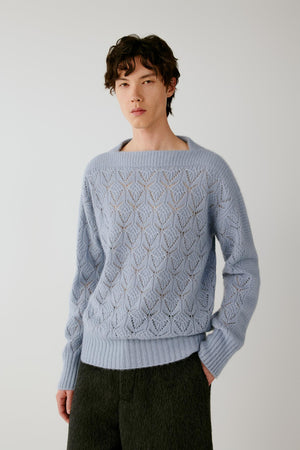 A man wearing a light blue knit sweater with a textured geometric pattern and dark charcoal pants, standing against a plain light gray background.