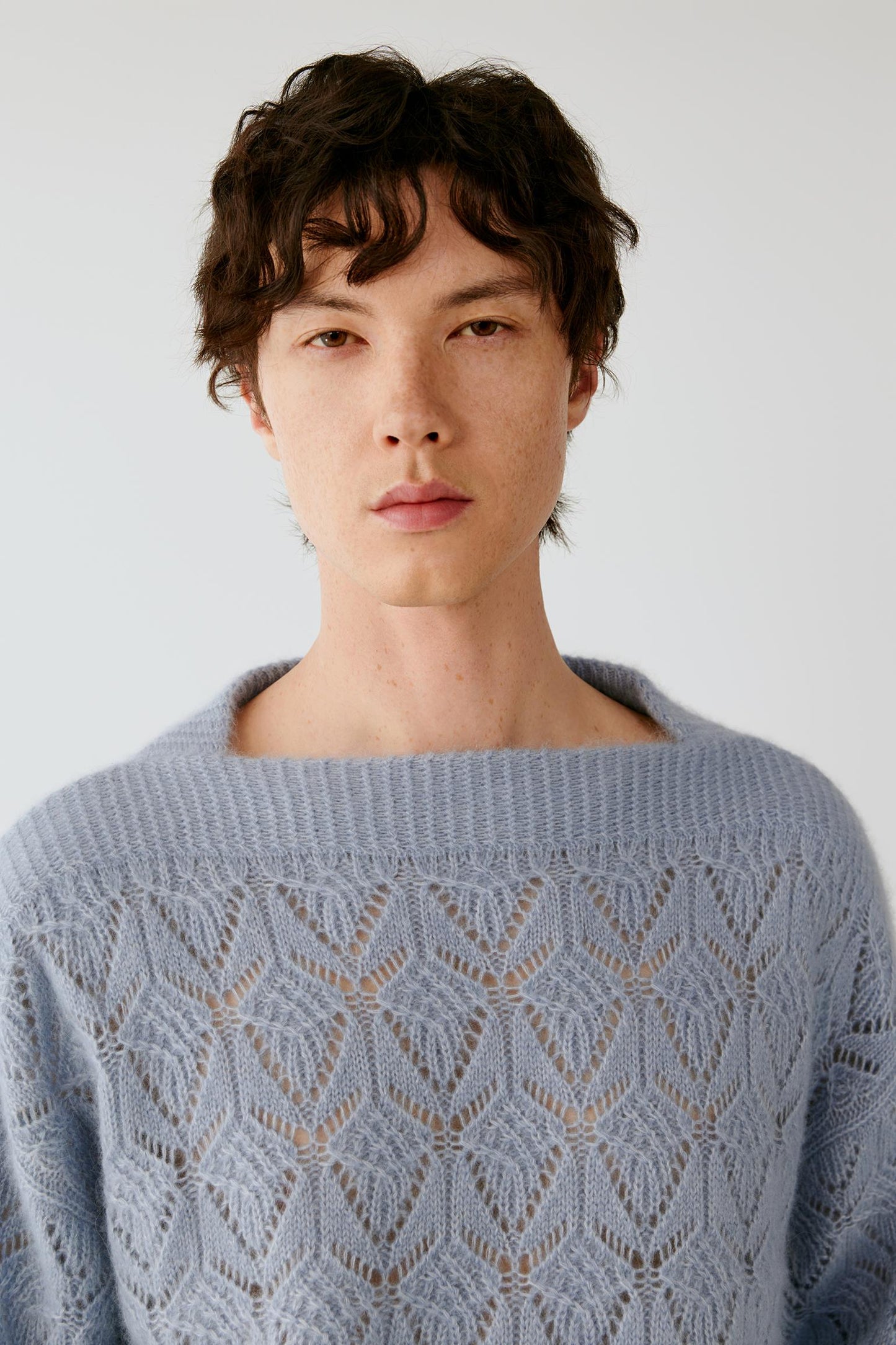 A man wearing a light blue open-knit crewneck sweater, photographed against a plain light gray background.
