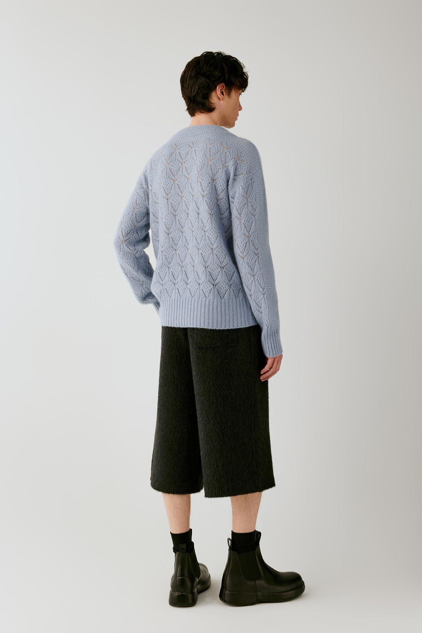 A model shown from the back wearing a light blue cable-knit sweater, dark charcoal cropped trousers, black socks, and black boots, against a plain light gray studio background.
