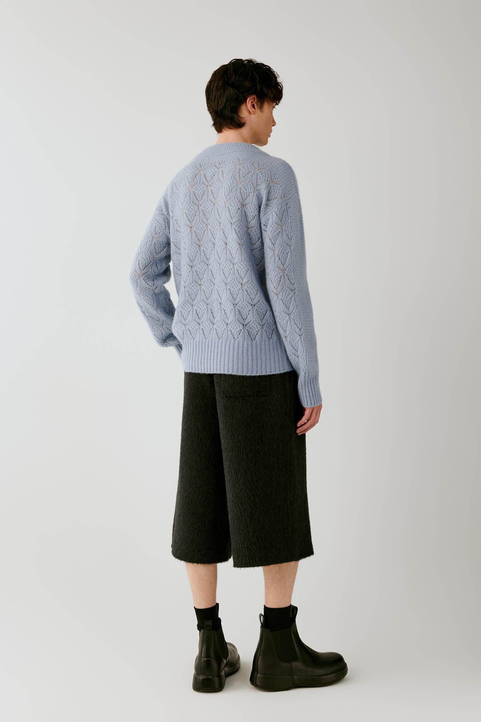 A model shown from the back wearing a light blue cable-knit sweater, dark charcoal cropped trousers, black socks, and black boots, against a plain light gray studio background.