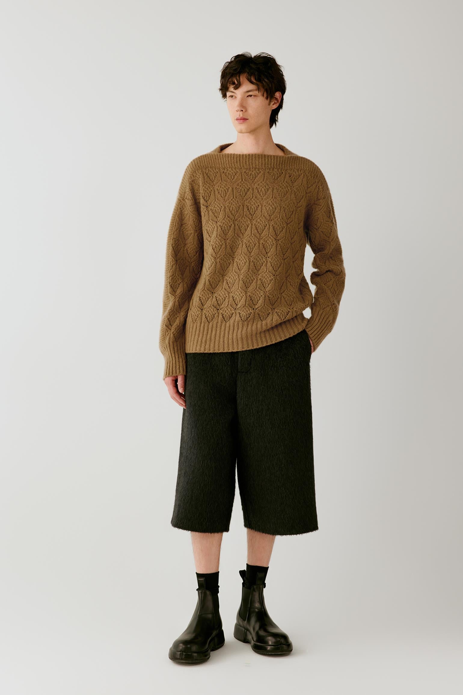 A model stands against a light gray studio background wearing a camel brown cable-knit sweater, dark charcoal cropped trousers, and black boots.