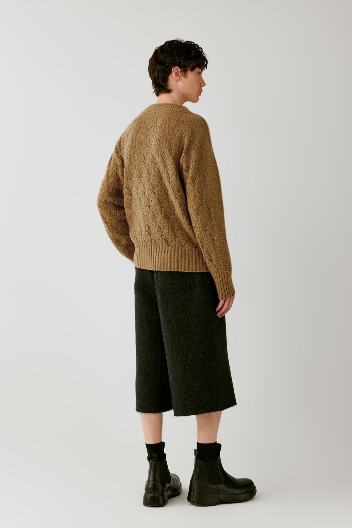 A model shown from the back wearing a tan cable-knit sweater, dark olive green cropped trousers, black socks, and black boots, photographed against a neutral light gray studio background.