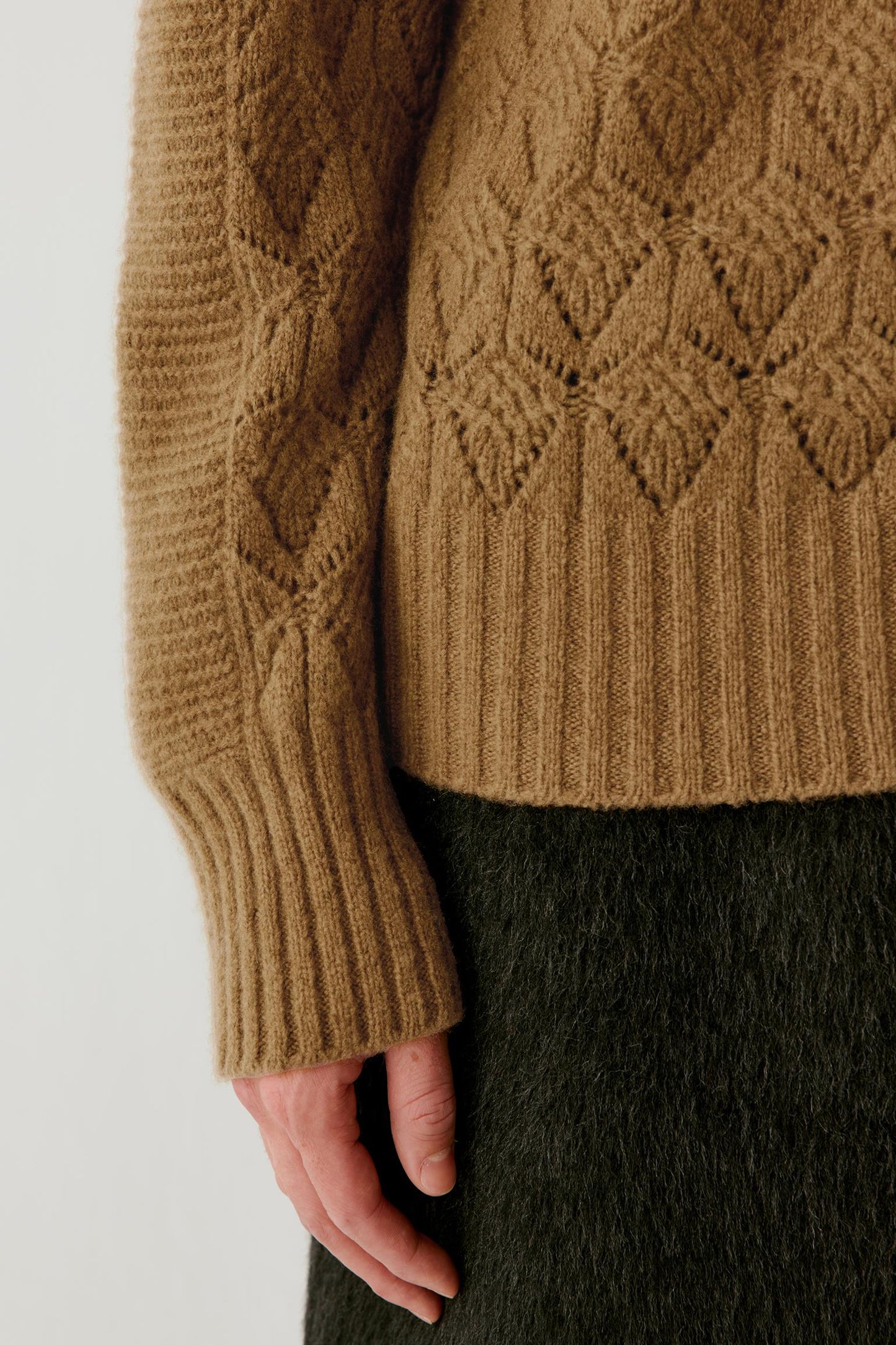 Close-up of a person wearing a chunky knit sweater in camel brown, showing the textured cable pattern and ribbed cuffs, paired with a dark olive skirt against a plain light background.