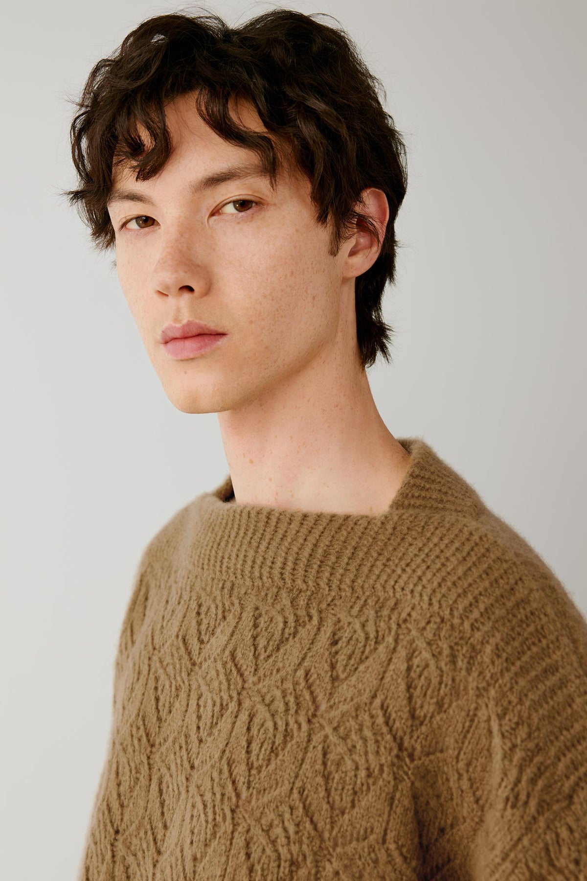 A man wearing a brown cable-knit crewneck sweater, photographed from the chest up against a plain light gray studio background.