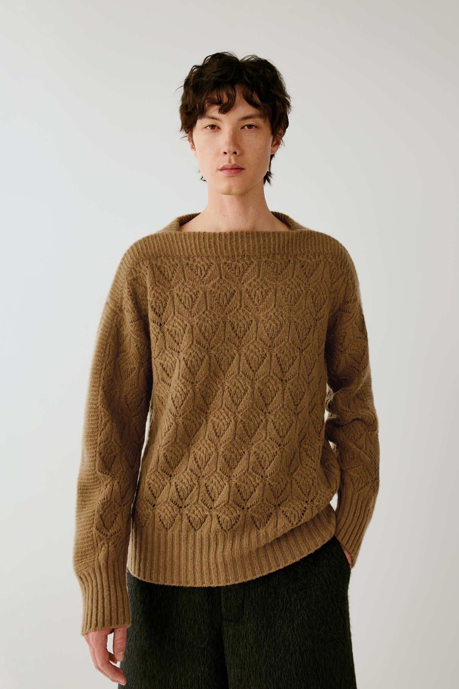 A man wearing a brown cable-knit sweater with a wide boat neckline, paired with dark green trousers, photographed against a light gray studio background.