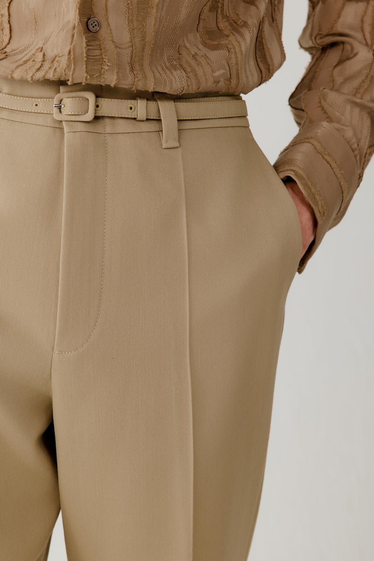 Close-up of a person wearing beige tailored trousers with a matching belt and a textured tan top, shown against a plain light background.
