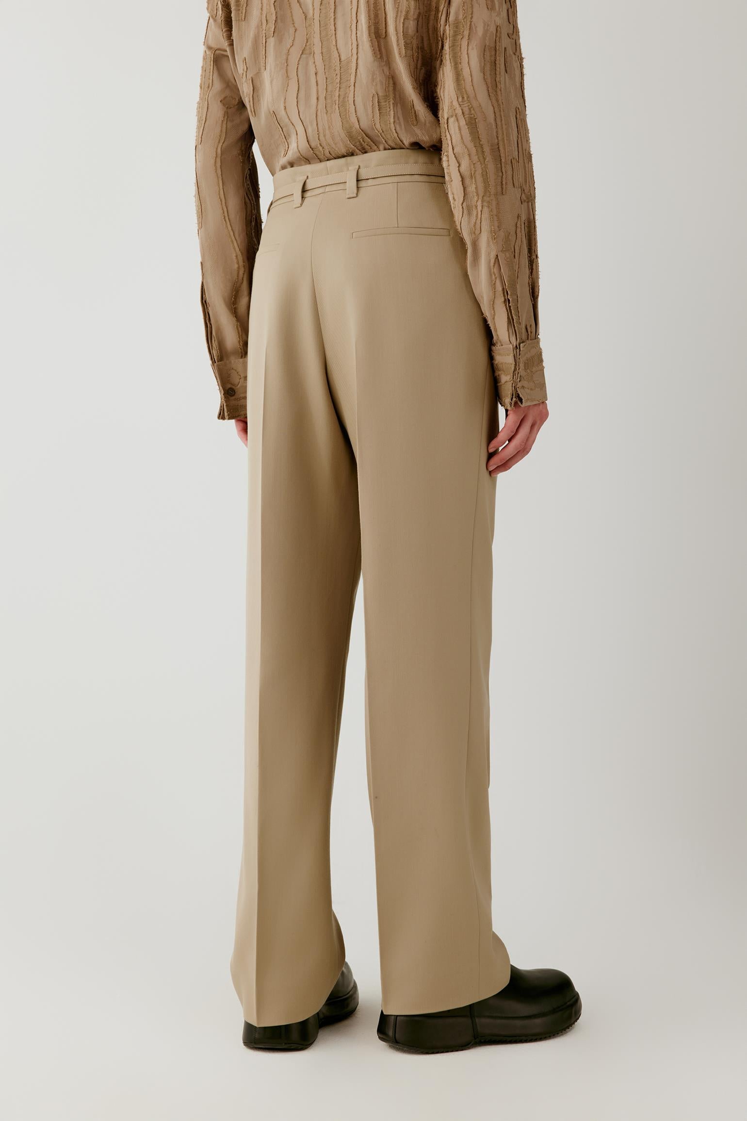 A person shown from the back wearing beige wide-leg trousers, a textured brown top, and black shoes against a plain light gray studio background.