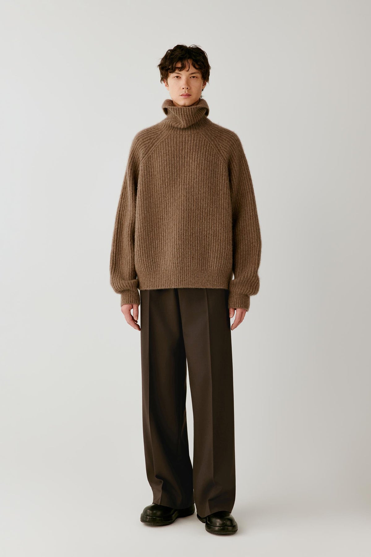 A model stands against a light gray studio background wearing an oversized ribbed knit turtleneck sweater in taupe-brown, paired with dark brown wide-leg trousers and black shoes.