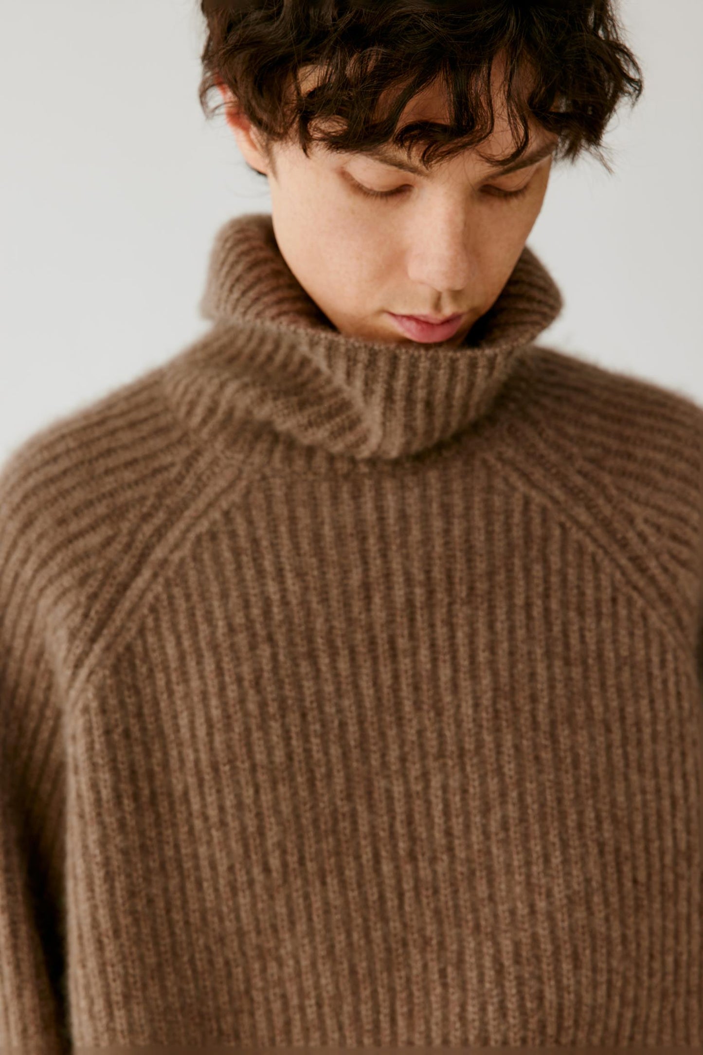 A close-up of a person wearing a brown ribbed knit turtleneck sweater against a plain light background.