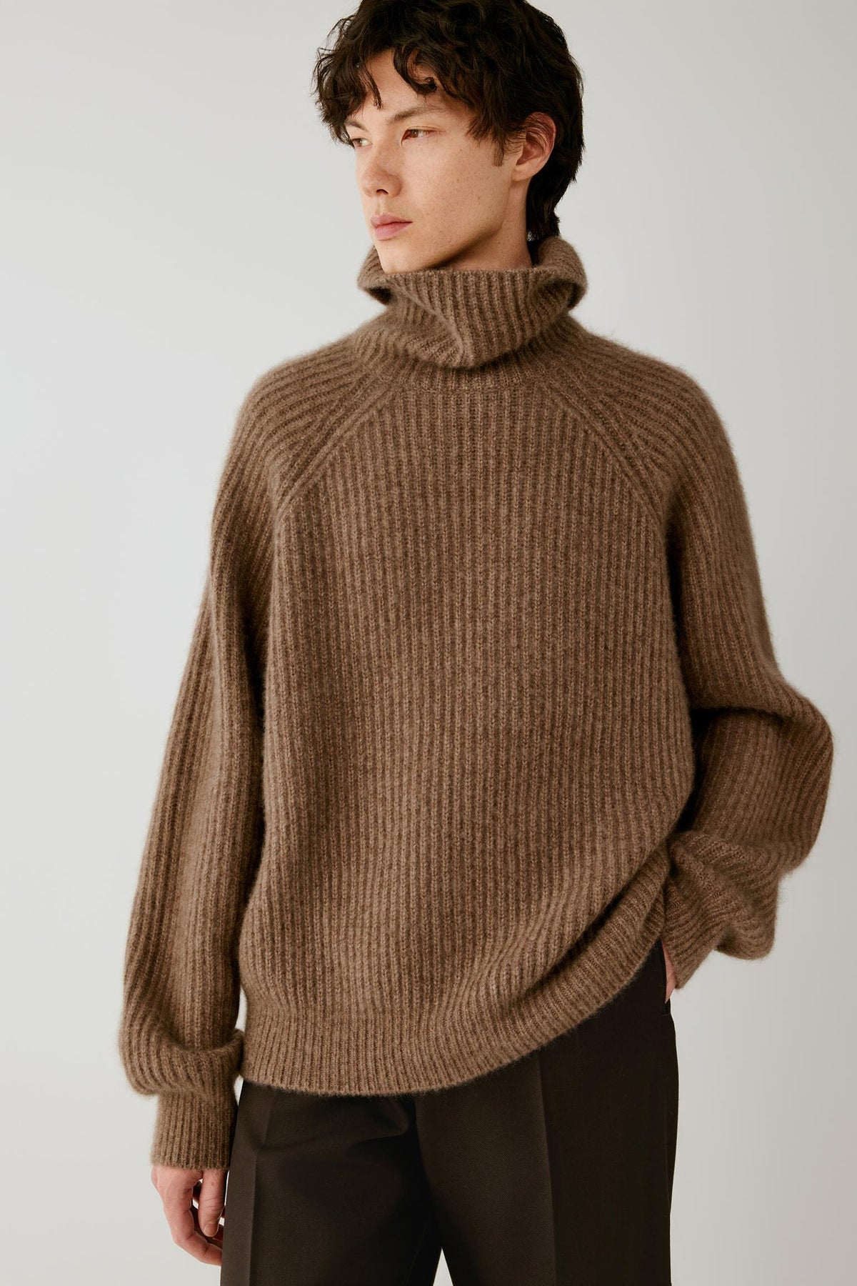 A man wearing a brown ribbed knit turtleneck sweater and dark trousers, photographed against a plain light gray studio background.
