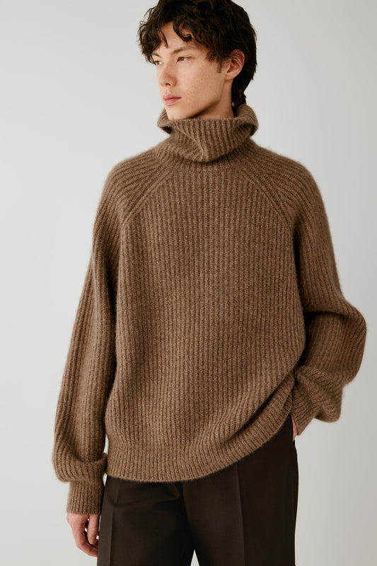 A man wearing a brown ribbed knit turtleneck sweater and dark trousers, photographed against a plain light gray studio background.