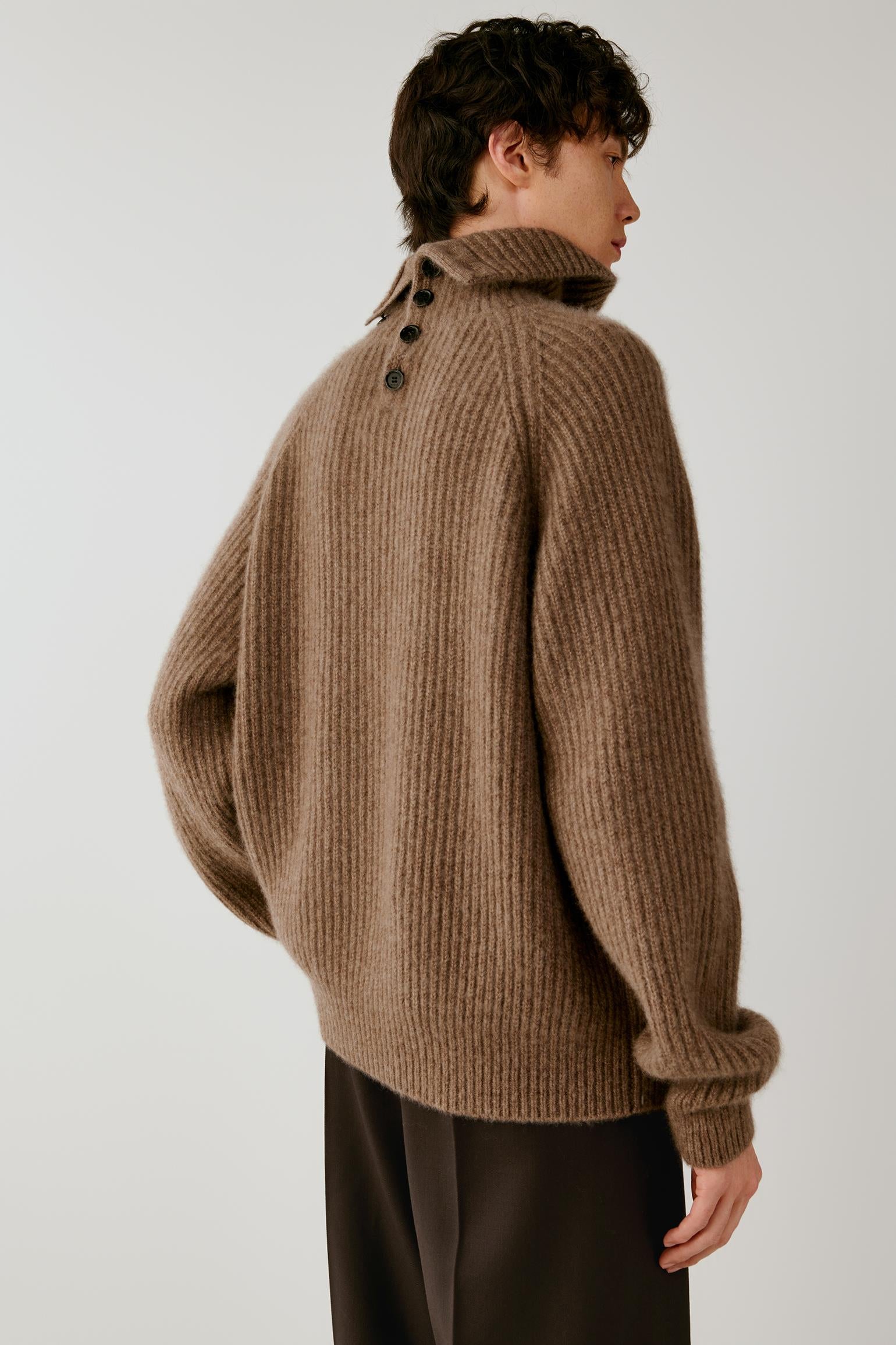 A man wearing a brown ribbed knit sweater with a high mock neck and dark buttons at the back, paired with dark trousers, photographed against a light gray studio background.