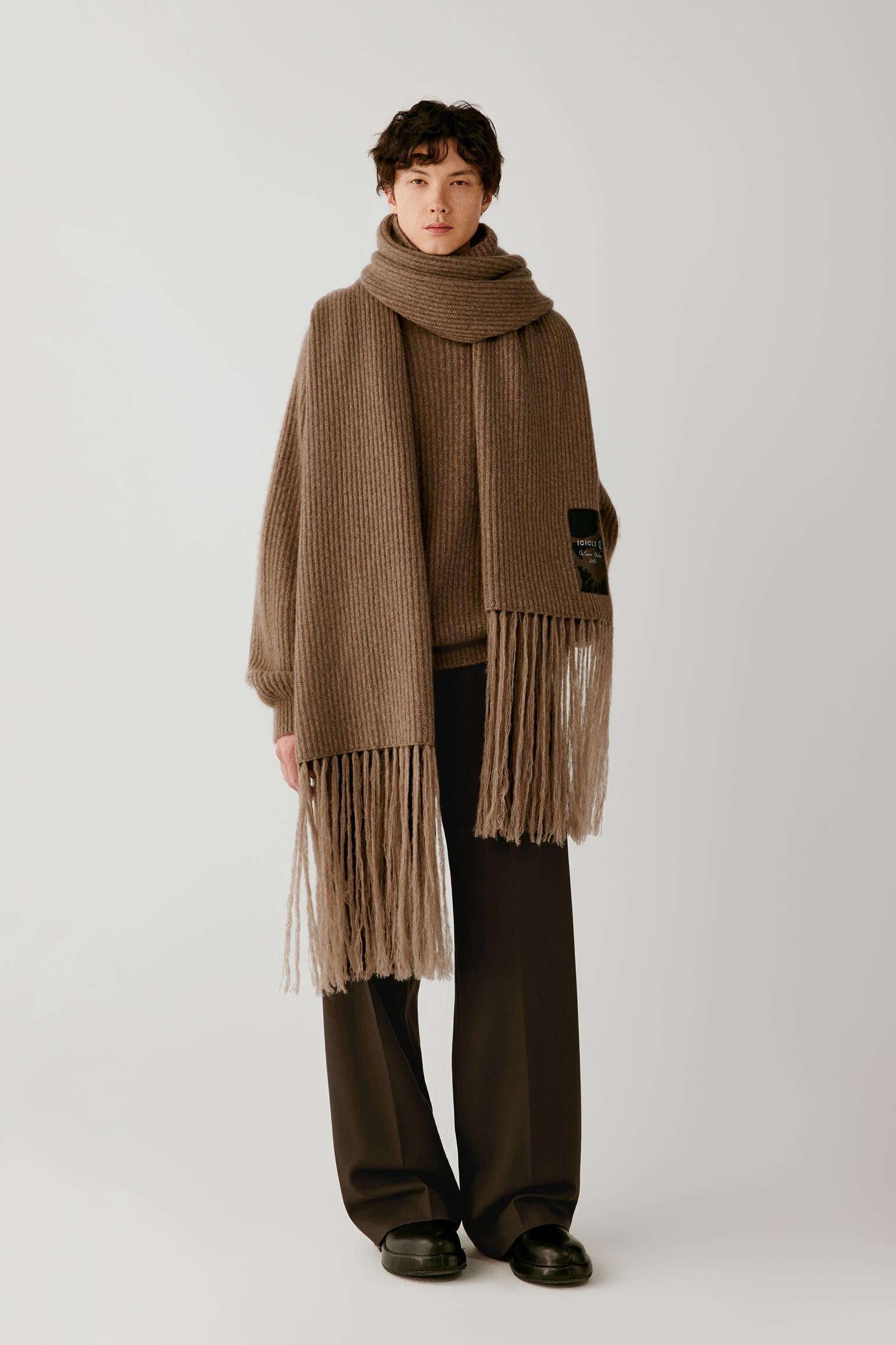 A model stands against a light gray studio background wearing a brown ribbed knit wrap scarf with long fringe, styled over a dark brown top and wide-leg dark brown trousers.