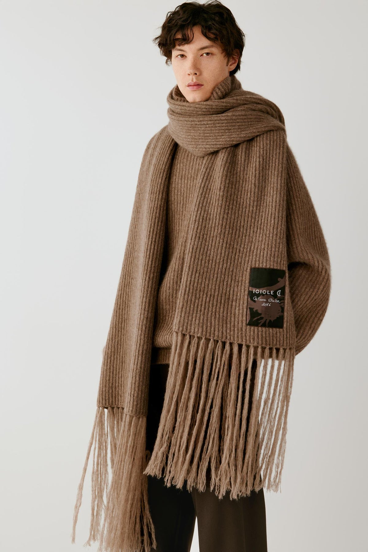 A model wearing a chunky ribbed knit brown scarf with long fringe and a matching brown sweater, photographed against a plain light gray studio background.