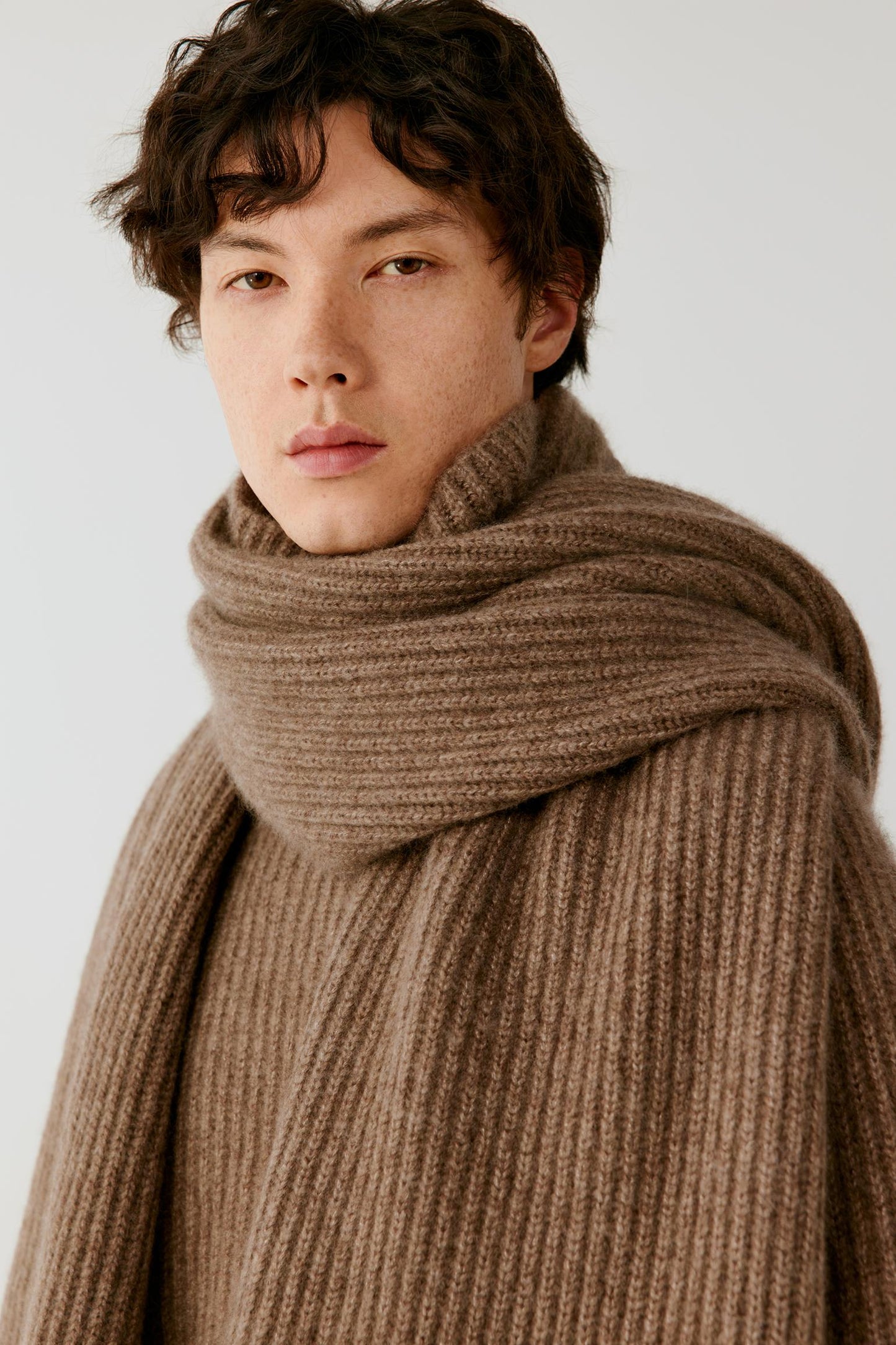 A model stands against a light gray studio background wearing a brown ribbed knit wrap scarf with long fringe, styled over a dark brown top and wide-leg dark brown trousers.