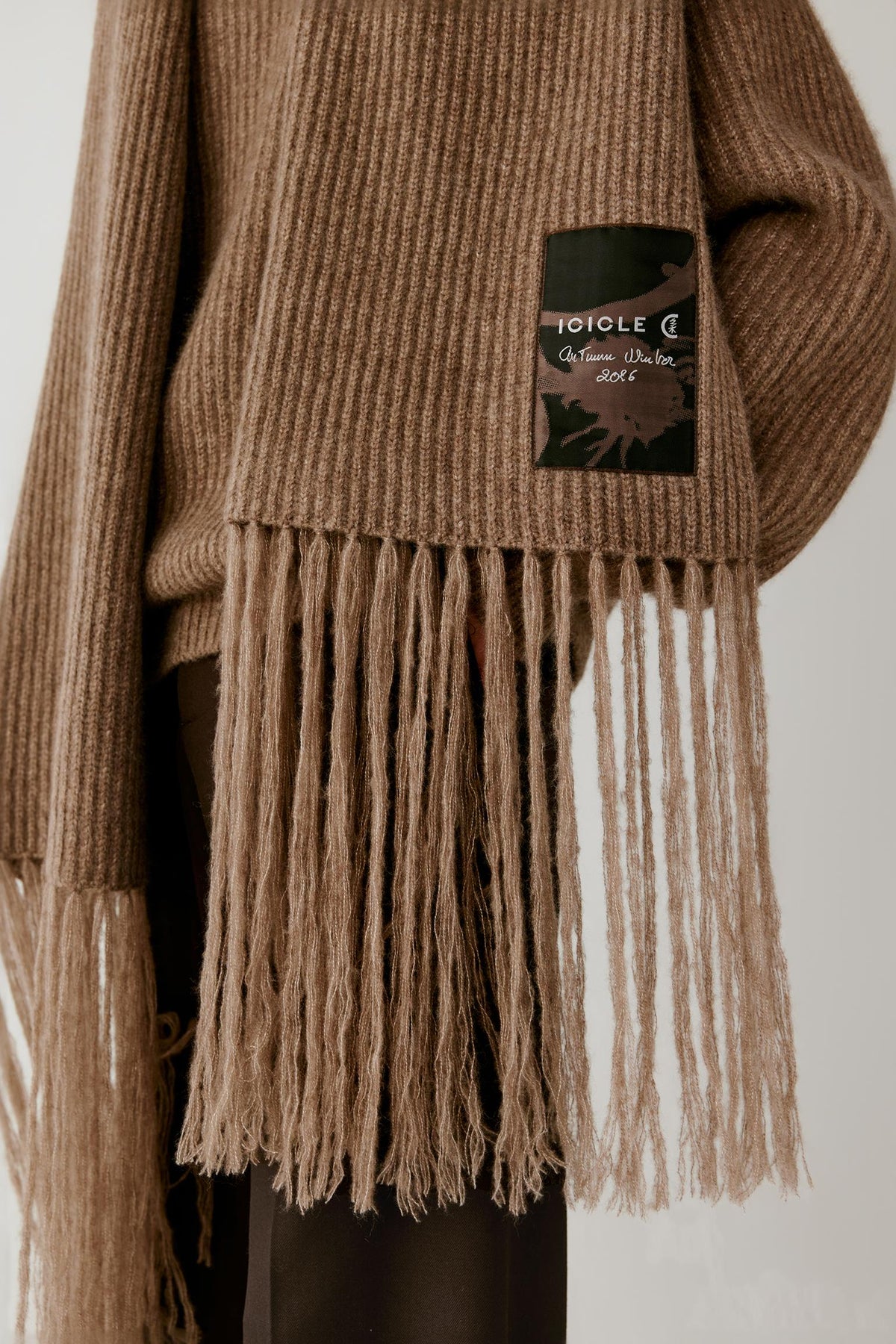 Close-up of a ribbed knit sweater in a warm taupe-brown color, featuring a long fringe hem and a black brand patch on the sleeve with the text 'JOULE C Collection Cotonnet'.