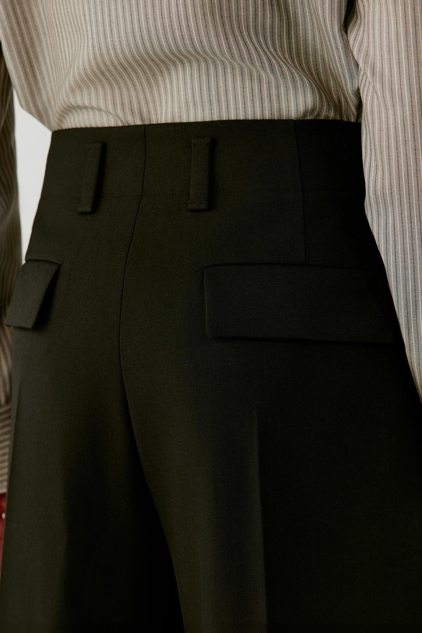 Close-up highlighting the waistband, belt loops and front pleat construction of the deep black wool twill trousers.
