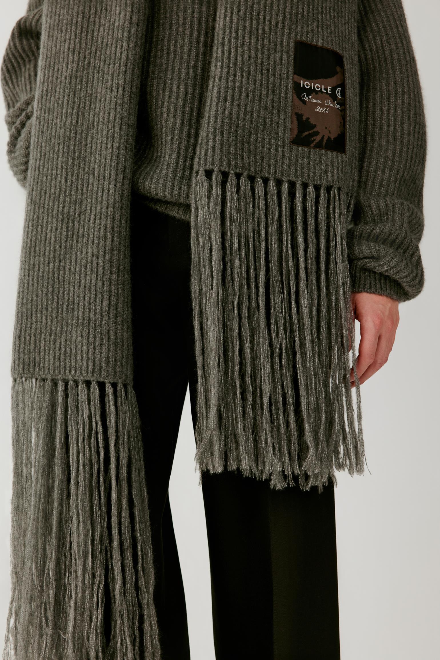 Close-up of a person wearing a ribbed knit sweater in a muted gray-green color with long fringe hanging from the hem and sleeve. A small black brand patch is visible on the upper chest, and the person is also wearing black pants against a plain light gray studio background.