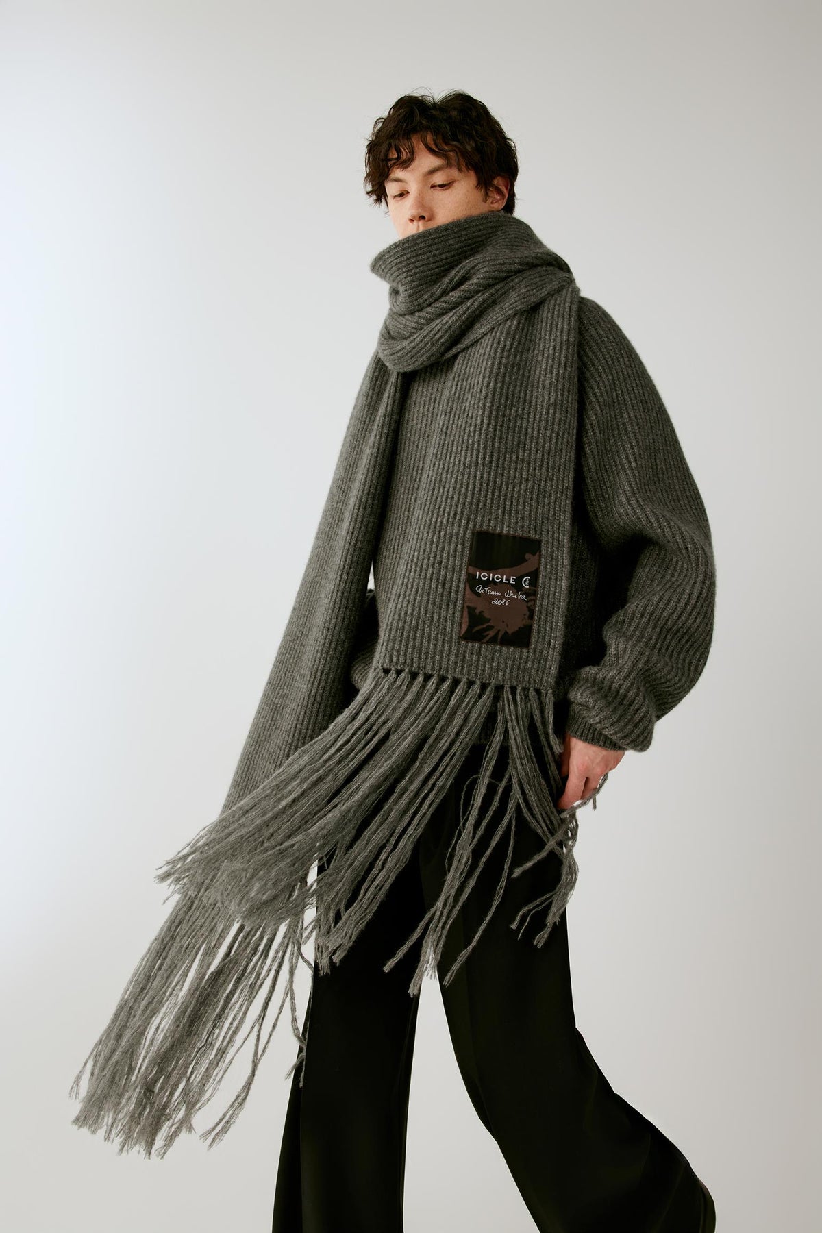 A model wearing a dark gray ribbed knit scarf with long fringe and a matching oversized knit sweater, standing against a light gray studio background.