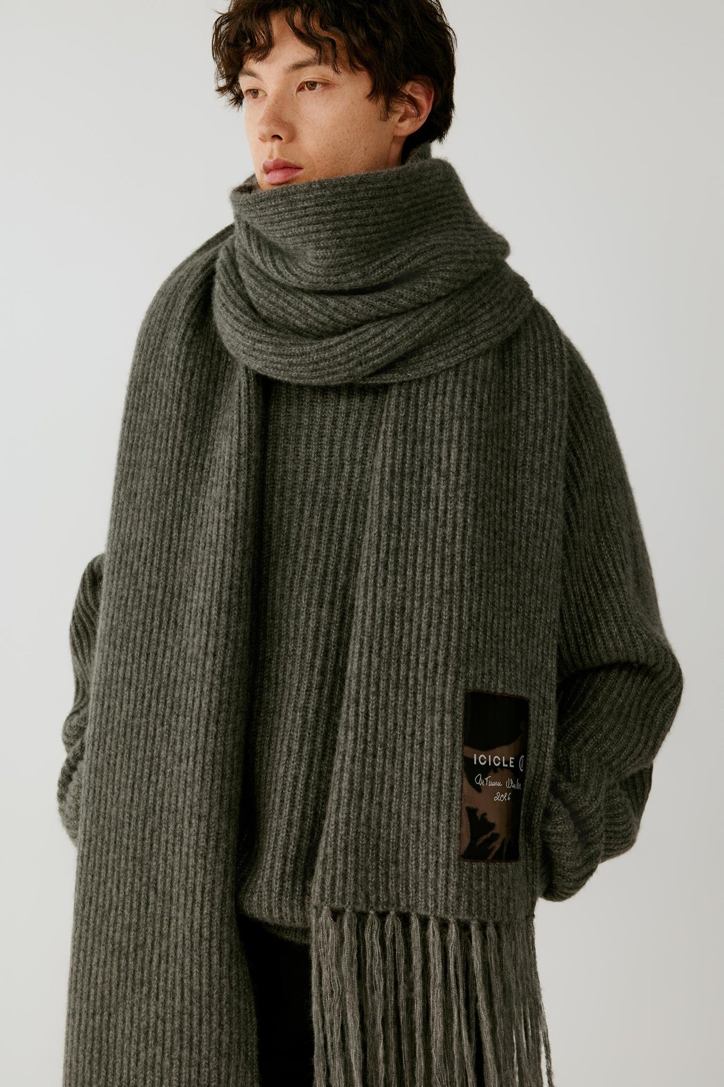 A model wearing a thick, ribbed knit scarf and matching oversized sweater in a dark gray-green color, featuring a small black brand patch on the front.