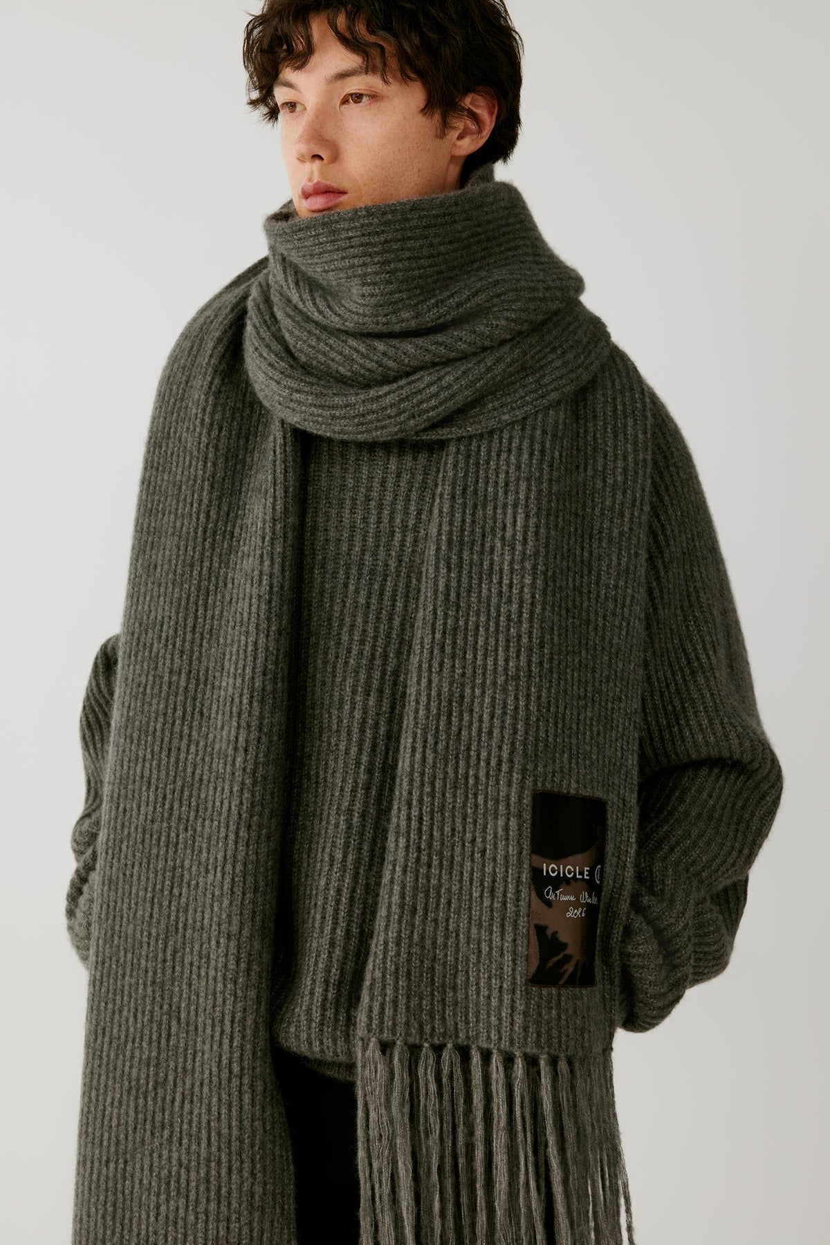 A model wearing a thick, ribbed knit scarf and matching oversized sweater in a dark gray-green color, featuring a small black brand patch on the front.