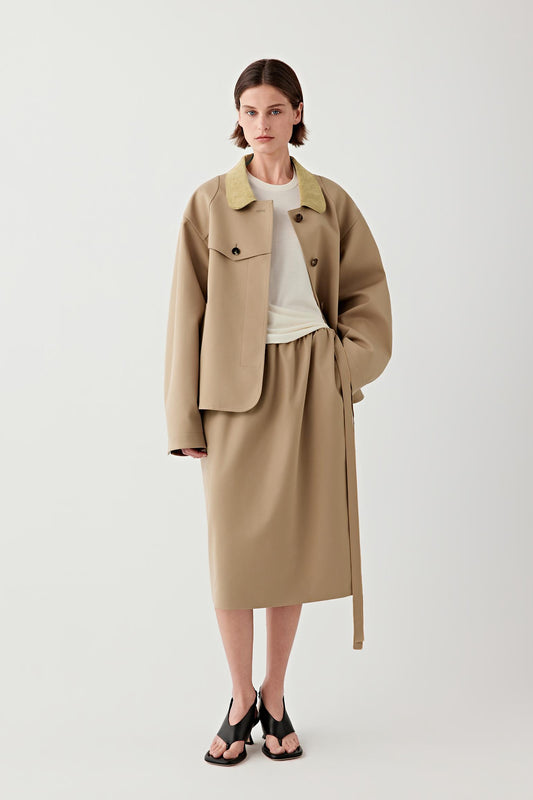 Woman wearing a short camel wool and silk trench coat with a relaxed, slightly oversized cut, styled with a matching skirt, full-length front view showing the soft structure, button fastening and belt detail