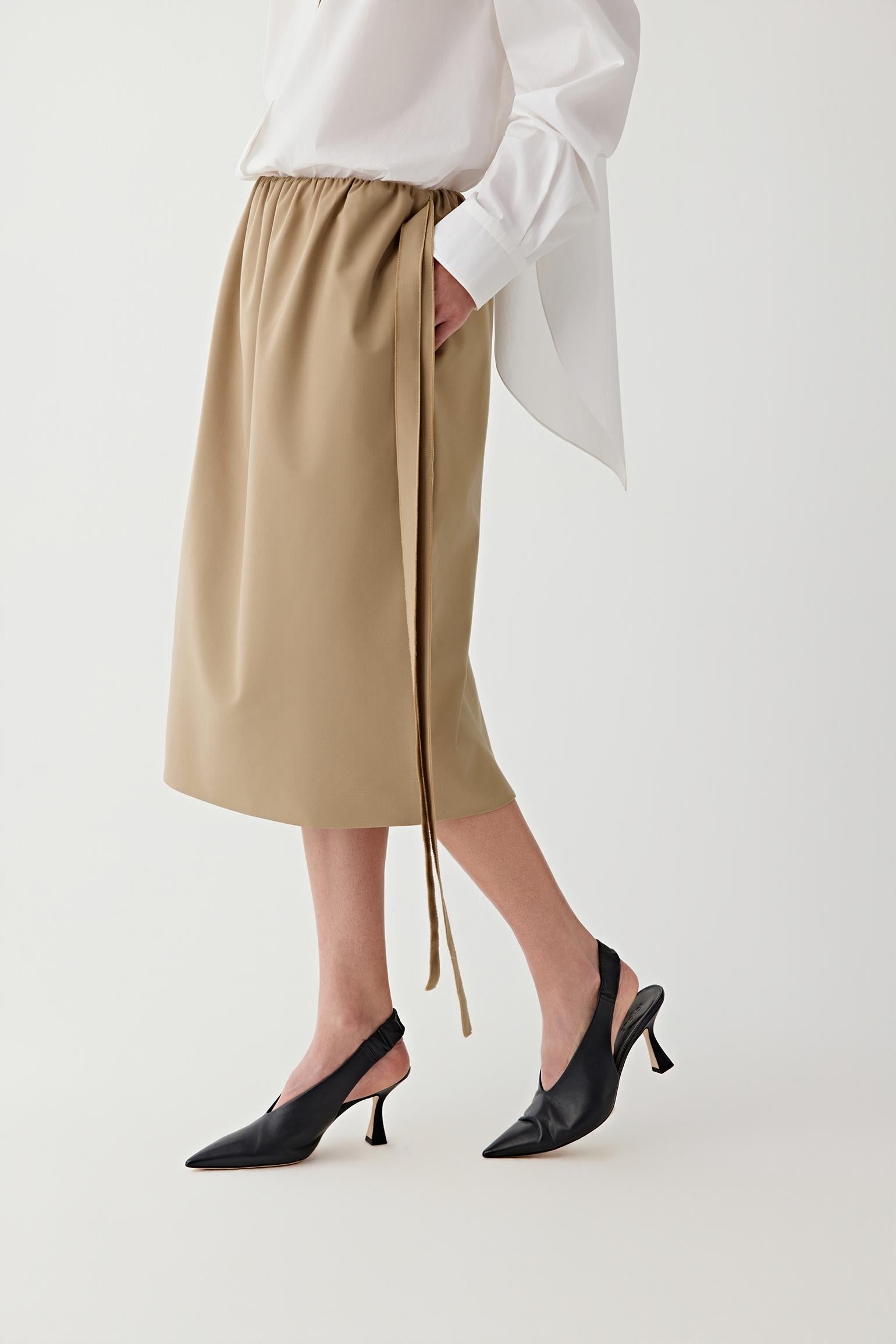 Person wearing a beige skirt with a white blouse and black shoes on a light background