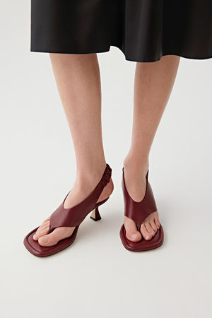 Close-up front three-quarter view of a model’s feet wearing deep red nappa leather heeled sandals, shown from the front, highlighting the square padded sole, toe post construction and wide front strap, with a smooth matte leather surface and rich, even colour