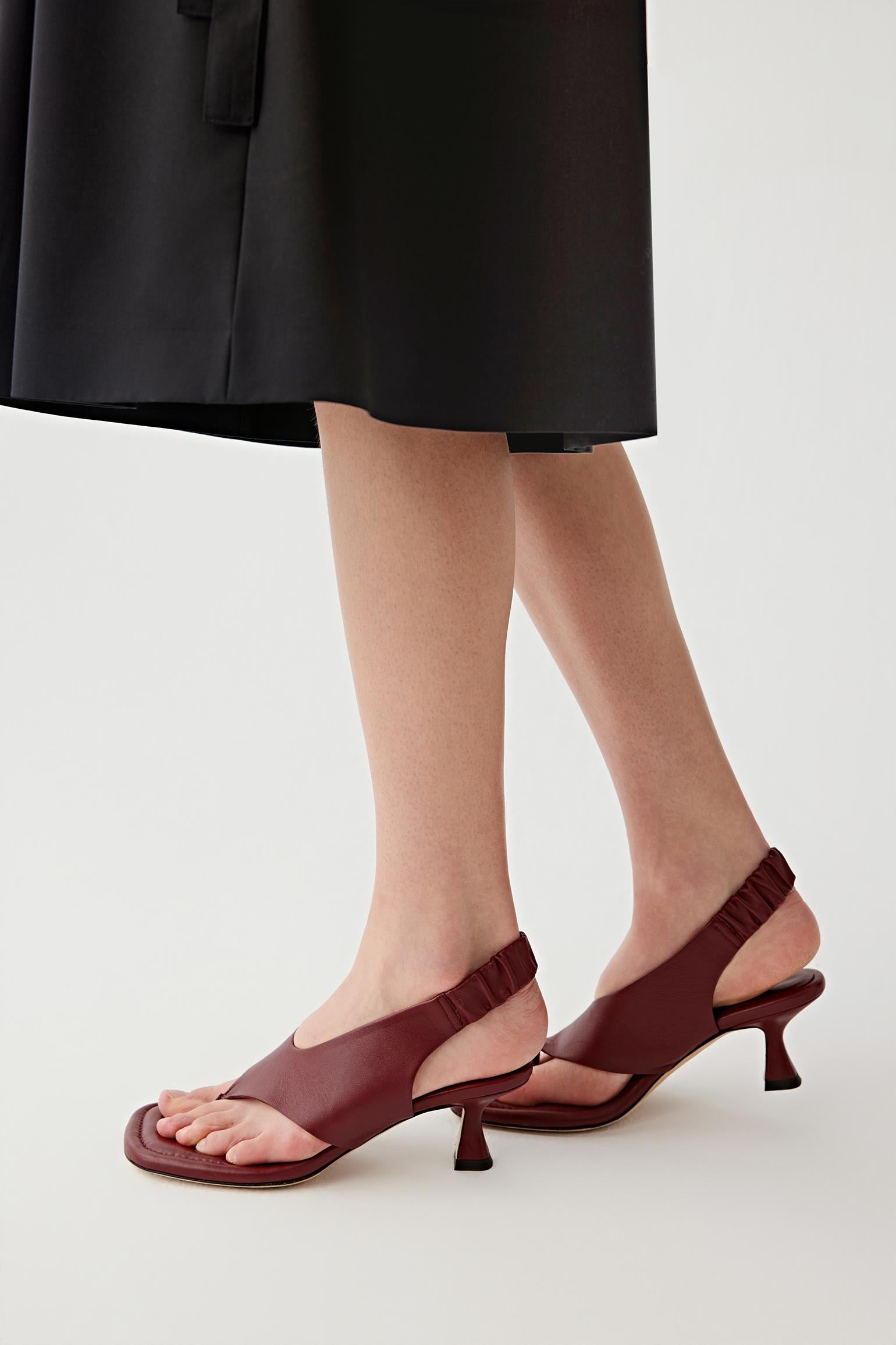 Side view of both feet showing the sandals worn in motion stance, emphasizing the curved mid heel, the slingback strap with gathered detail around the heel and the balanced structure between sole, upper and heel