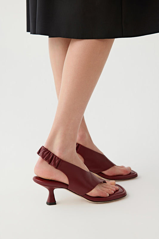 Side view of both feet showing the sandals worn in motion stance, emphasizing the curved mid heel, the slingback strap with gathered detail around the heel and the balanced structure between sole, upper and heel