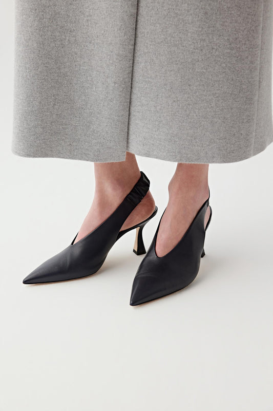 Close-up front three-quarter view of a model’s feet wearing deep black nappa leather slingback heels, paired with light grey trousers, highlighting the pointed toe shape, smooth matte leather surface and clean, elongated silhouette from toe to vamp