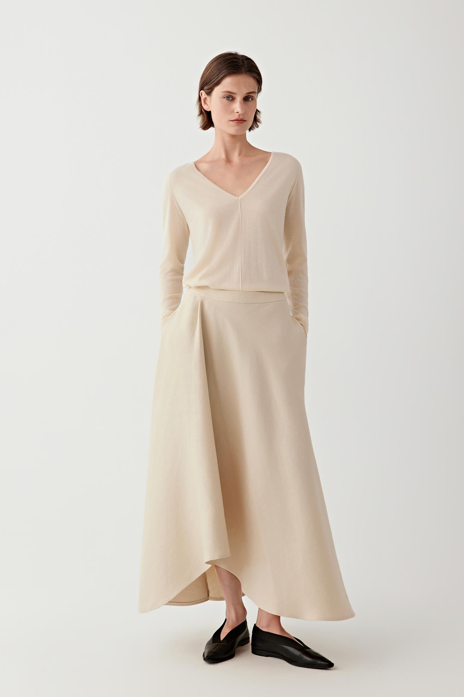 Female model wearing a beige set featuring a long-sleeve V-neck sweater and a fluid asymmetrical maxi skirt, paired with black loafers.