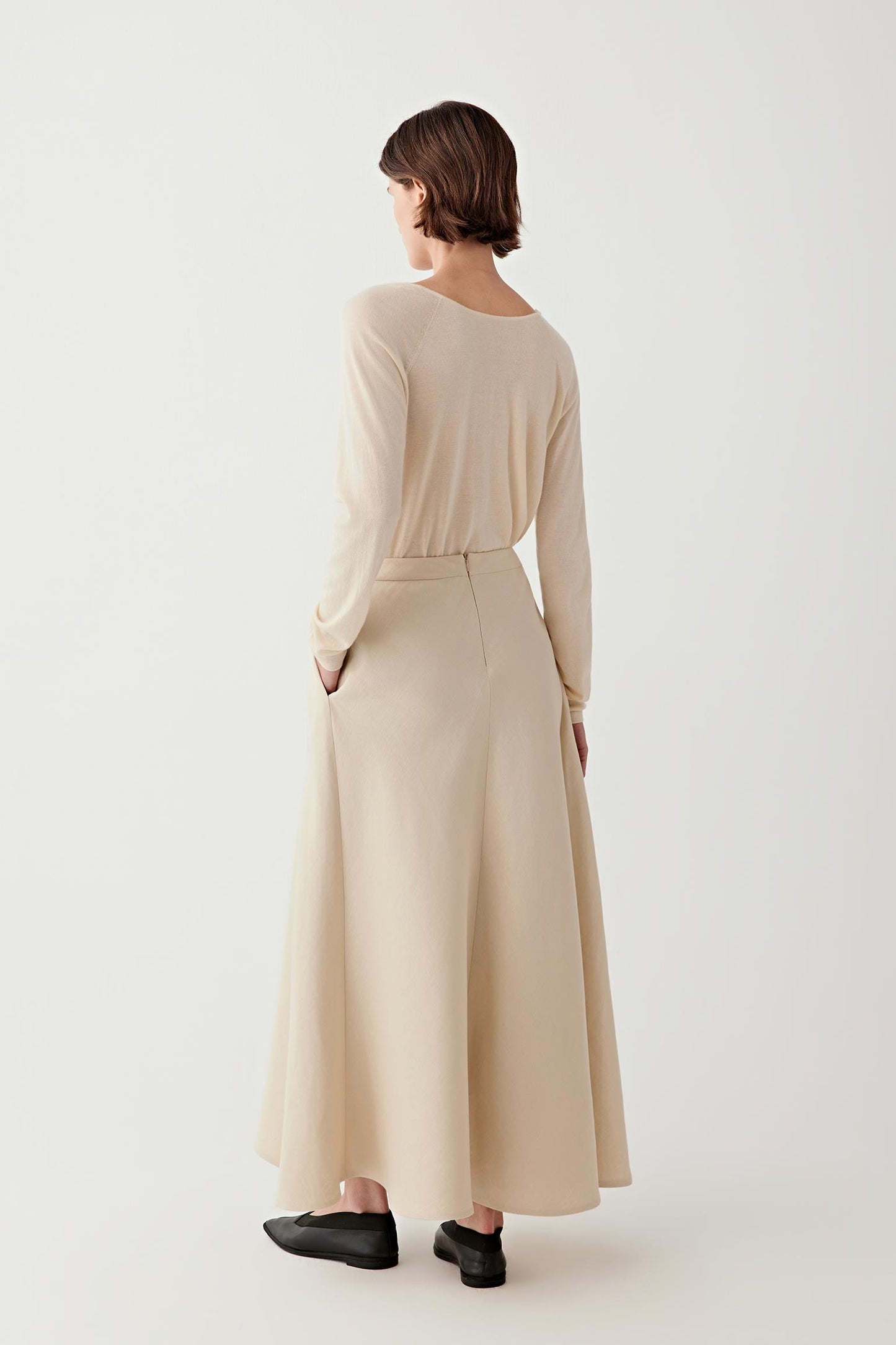 Back view of a female model wearing a beige long-sleeve knit top and a fluid maxi skirt, paired with black loafers.