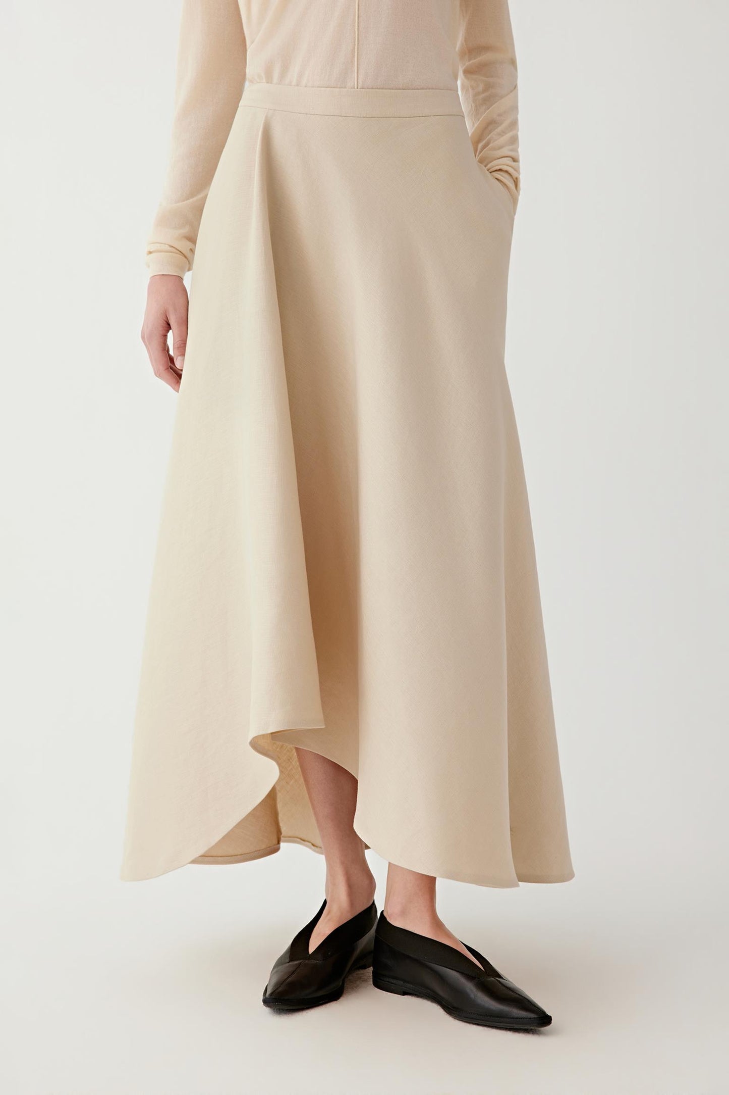 Detail of a beige maxi skirt with an asymmetrical cut and fluid drape, worn with black loafers.