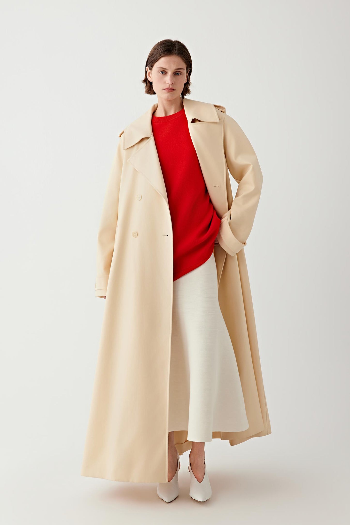 Woman wearing a beige coat over a red sweater and white skirt on a light background