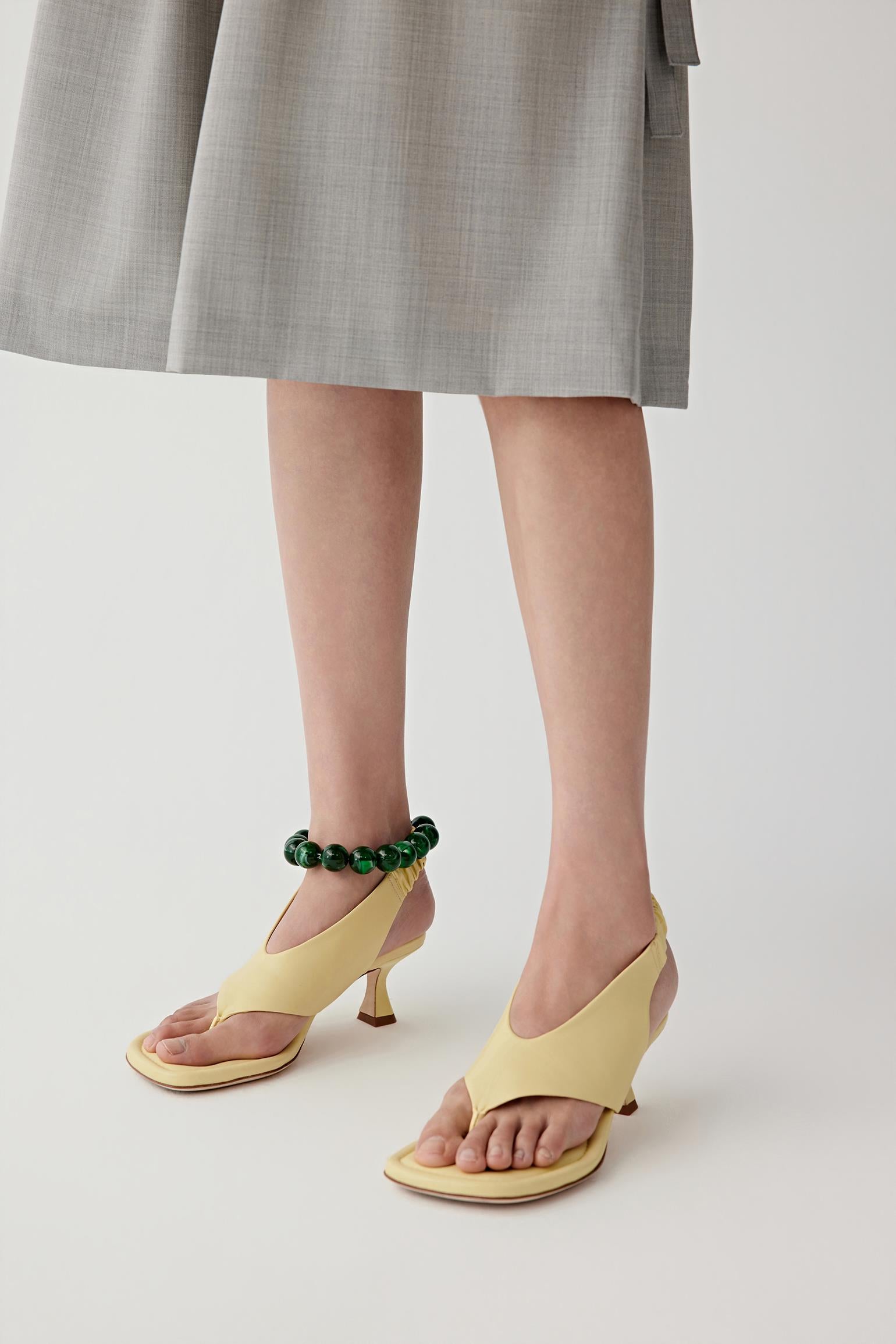 Close-up front three-quarter view of a model’s feet wearing pale yellow nappa leather heeled sandals, showing the square padded sole, toe post construction and wide front strap, with a smooth matte surface and soft leather finish