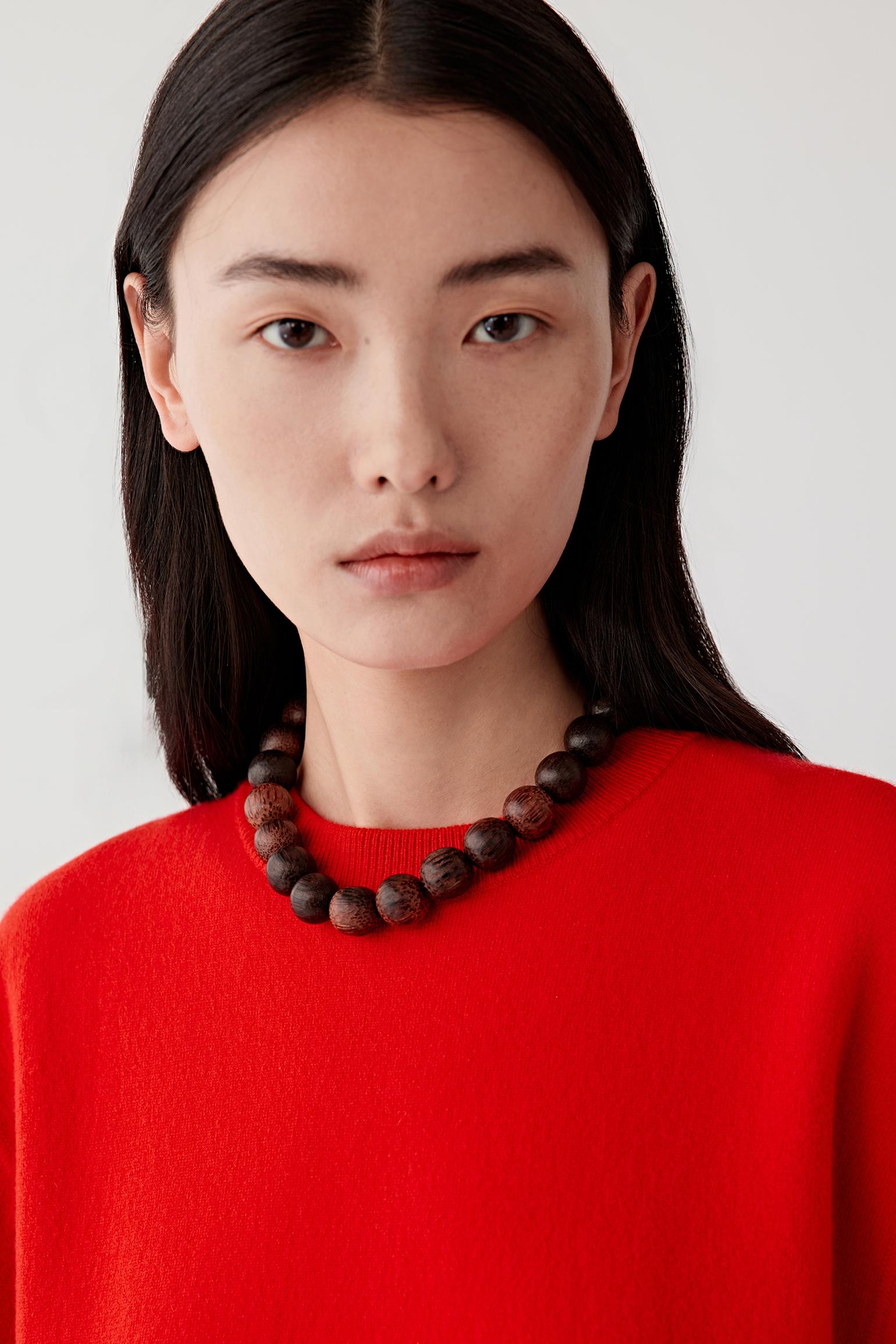Woman wearing a red sweater and wooden beaded necklace against a white background