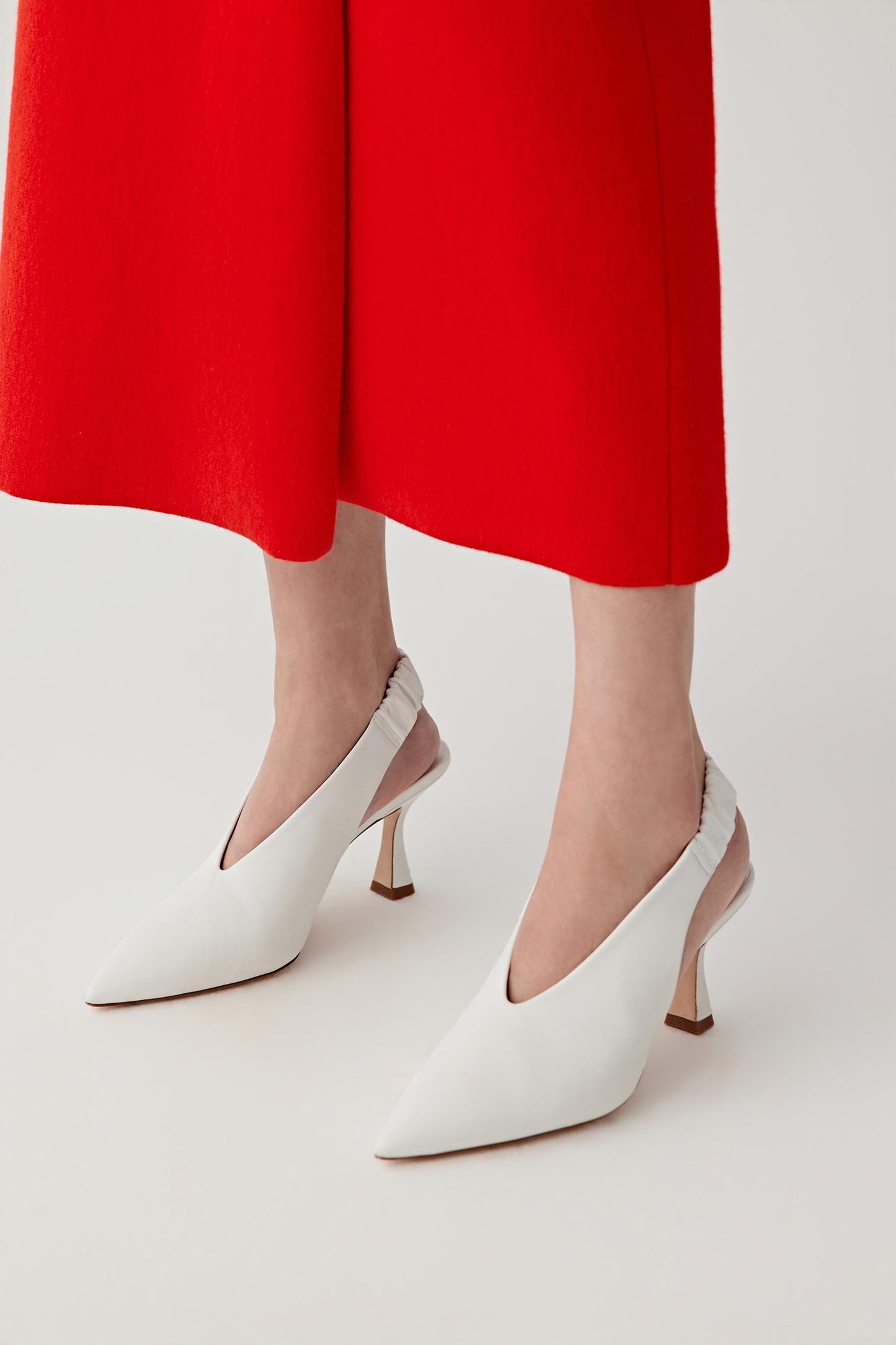 Close-up front three-quarter view of a model’s feet wearing white nappa leather slingback heels, paired with a red skirt, highlighting the pointed toe shape, smooth matte leather surface and clean, elongated front line