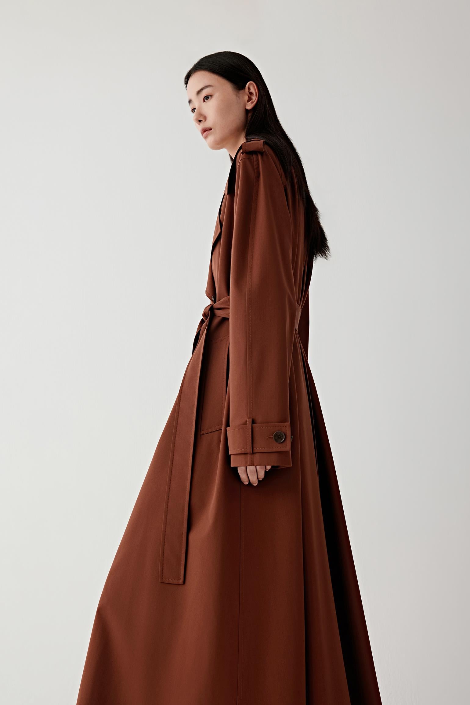 Three-quarter view of a woman wearing a long brown trench coat in wool and silk with a wide collar and a belt tied at the waist, showing the structured shoulders and elongated silhouette.