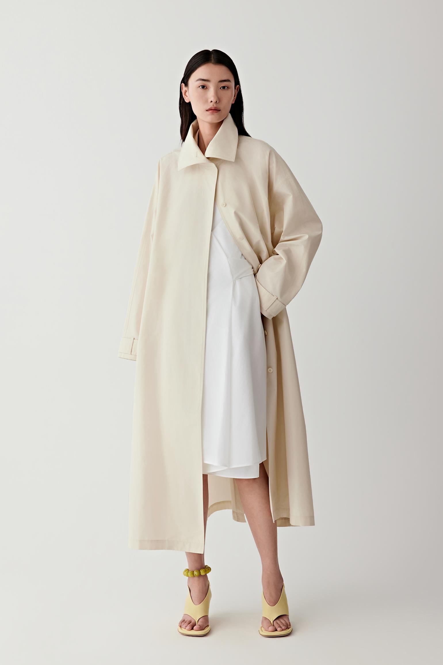Woman wearing a beige coat over a white dress on a white background