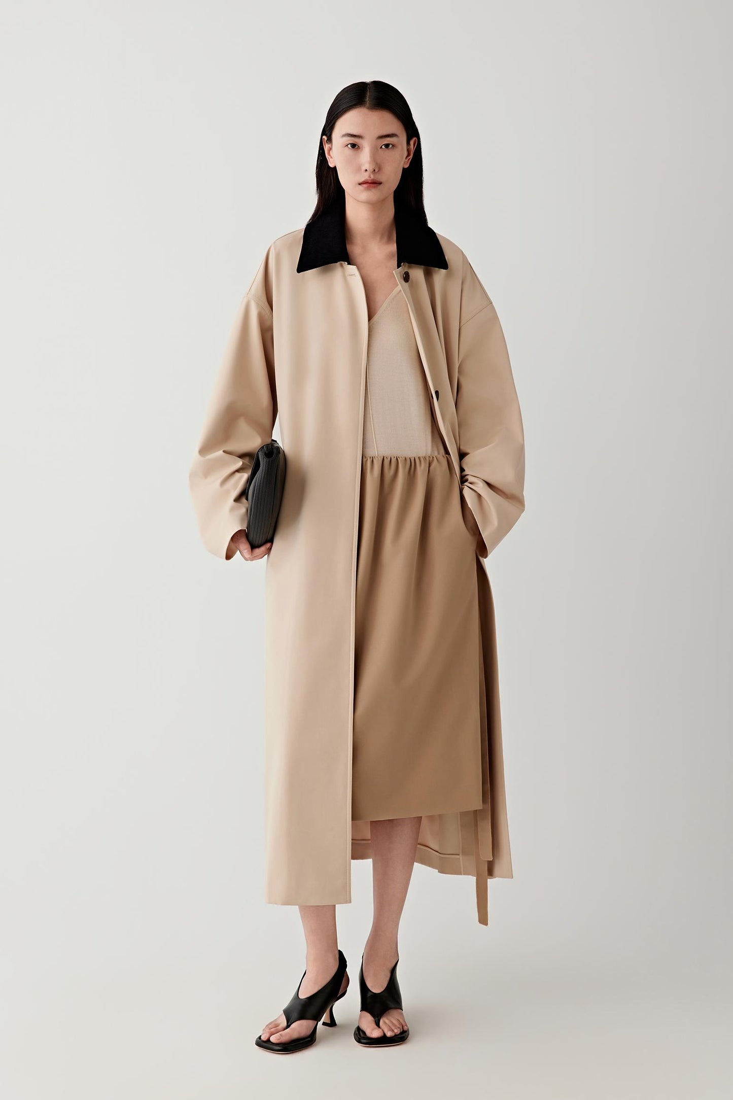 Woman wearing a beige coat and dress holding a black clutch against a white background