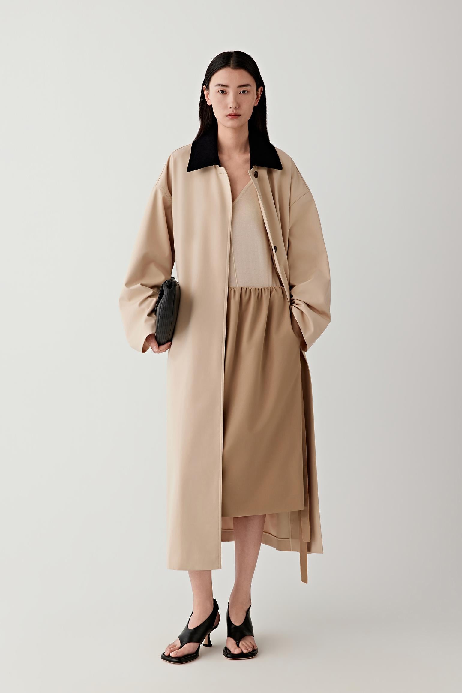 Full-length studio photograph of a woman wearing a long beige DEW trench coat in wool and silk with a removable contrasting dark collar. The coat has a straight elongated silhouette, relaxed shoulders and wide sleeves, falling below the calf.