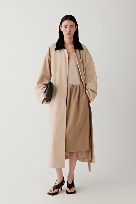 Woman wearing a beige coat and dress holding a black clutch against a white background