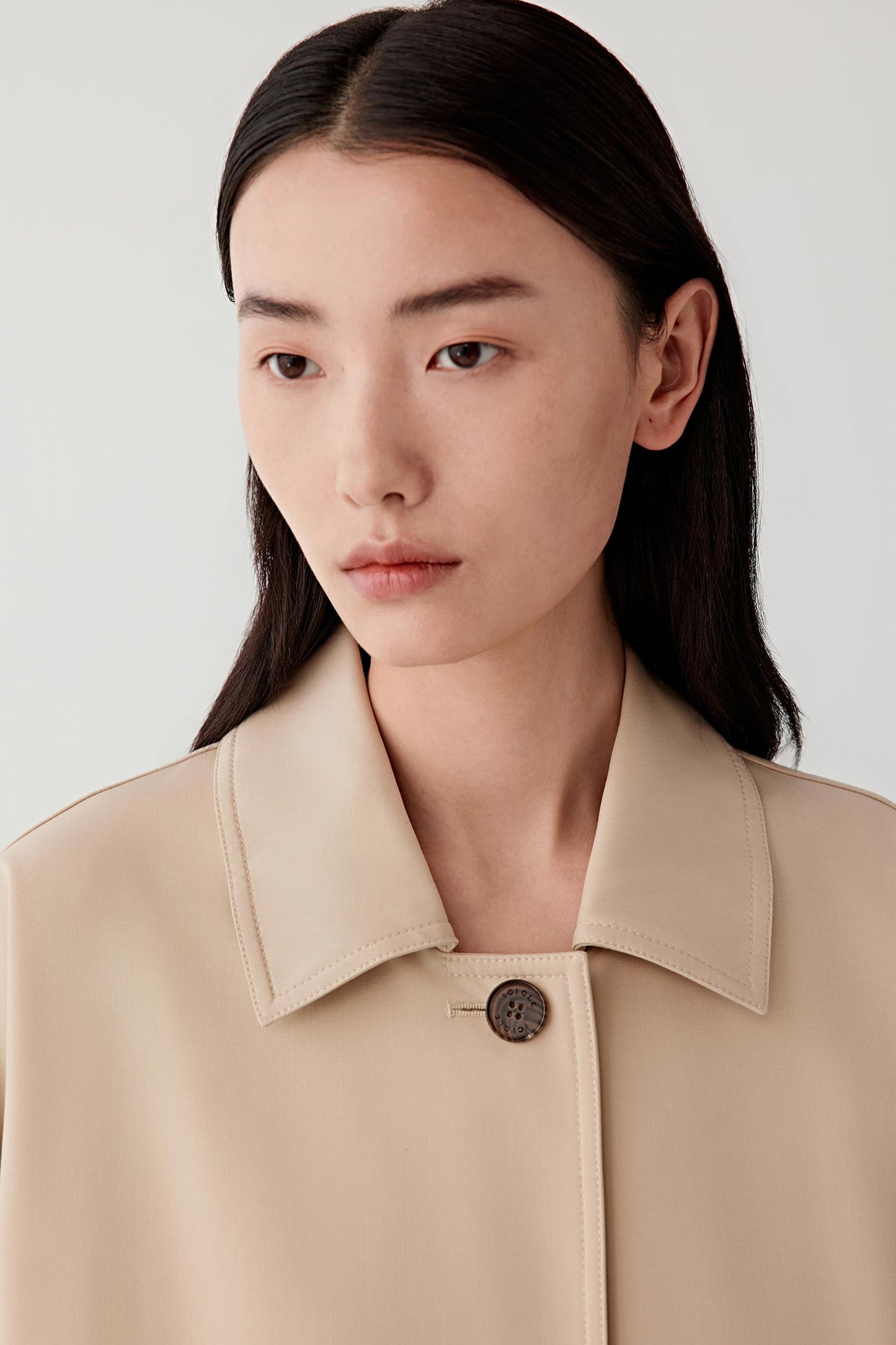 Close-up portrait of a woman wearing a beige jacket with a pointed collar and a single visible button, against a plain light background.