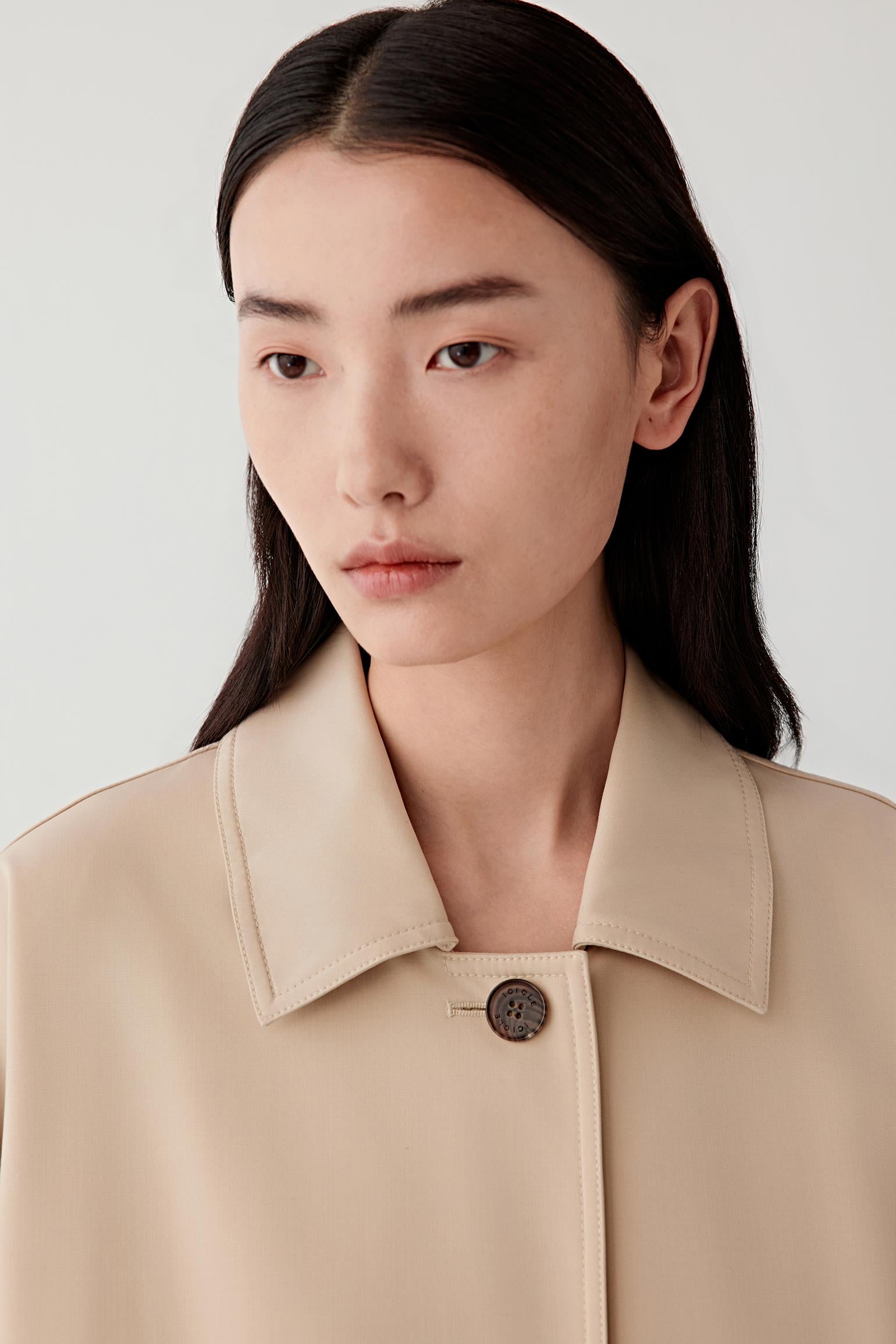 Close-up portrait of a woman wearing a beige jacket with a pointed collar and a single visible button, against a plain light background.