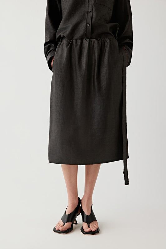 Front view of a woman wearing a liquorice-black midi skirt in Gambiered Canton Gauze, highlighting the textured matte surface, fluid drape and tonal depth created through the traditional dyeing process. The fabric reveals subtle irregularity and a softly structured appearance.
