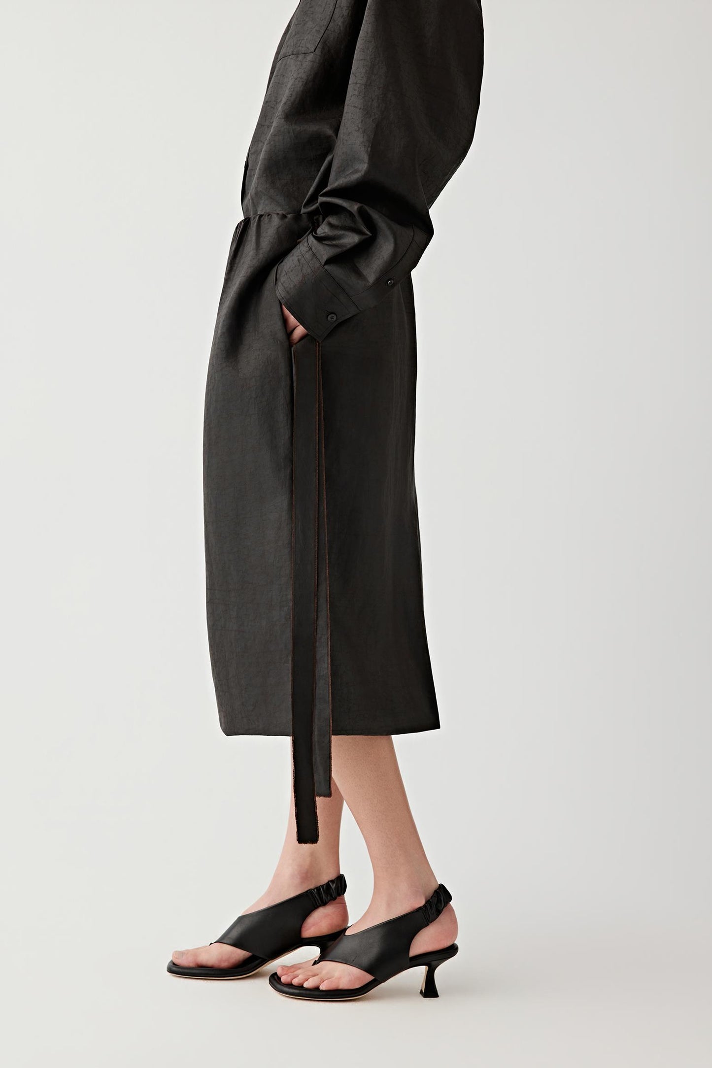 Side view of the sandals worn with a mid-length black skirt, emphasizing the curved mid heel, the slingback strap with gathered detail at the back of the ankle and the balanced proportion between sole, heel and upper