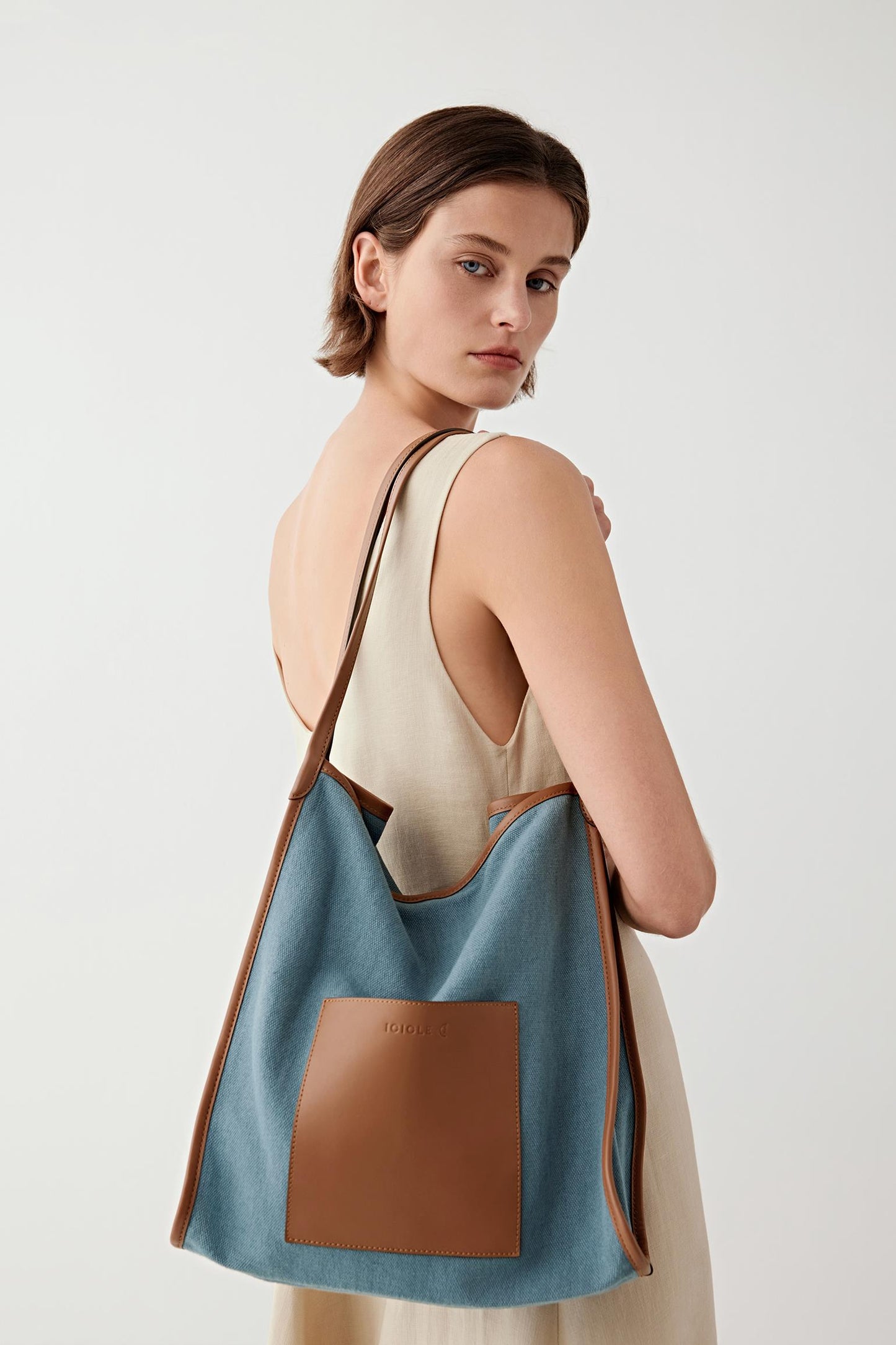 Back three-quarter view of a woman carrying a blue linen and cotton tote bag with brown leather panel, showing soft structure, shoulder carry and color contrast