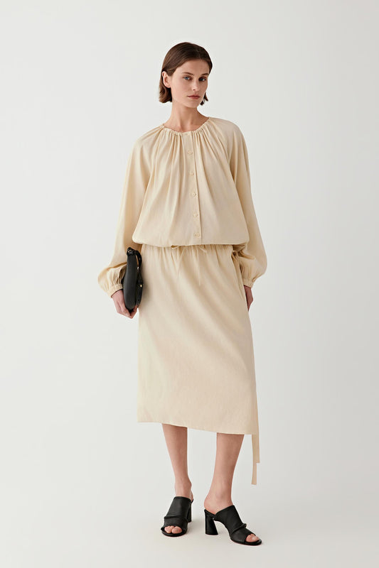 Woman wearing a sand-colored blouse with gathered neckline and long sleeves, paired with a matching skirt, full-length view.
